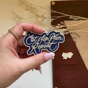 May include: A blue embroidered patch with white stitching that reads "Eat, Sip, Plan, Repeat".
