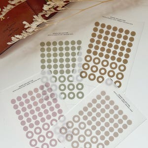 May include: Four sheets of clear sticker paper with different coloured dots and rings. The colours are olive green, light brown, dusty rose, and taupe.