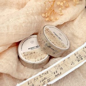 May include: Two rolls of Script No. 4 vintage series washi tape with handwritten script and numbers. A strip of the tape is unrolled, displaying more of the script. The tape is beige with black text. The background is a beige fabric.