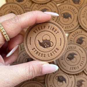 以下が含まれることがあります： 茶葉のデザインが施されたティーポットのグラフィックと「Steep First, Stress Later」というテキストが書かれた茶色の革製のコースターのクローズアップ。コースターは手で持たれています。