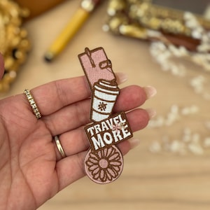 May include: A collection of embroidered patches showing a pink passport, a coffee cup, a sign reading "TRAVEL MORE", and a pink flower. The patches are brown and white with pink detailing.