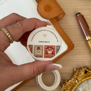 May include: A roll of washi tape with a coffee-themed design. The tape features postage stamp-style illustrations of coffee cups and coffee pots. The text "CAFE NO.3" is visible on the label. The tape is held in front of a notebook and pen.