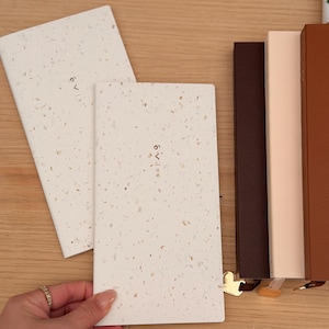 May include: Two cream notebooks with speckled patterns and Japanese text are held by a hand. Several other notebooks in shades of brown and tan are stacked nearby. Pens are visible in the background.