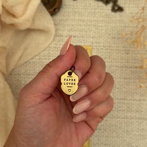May include: A small, shield-shaped brass charm featuring the text "PAPER LOVER" in black lettering, with a small heart symbol below the text and a jump ring at the top for attachment. The charm is held against a yellow ruler for scale.