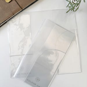 May include: Clear plastic menu holders with a curved top and a pocket. The text "KOLOUR CAFE" is printed on the front. A brown leather journal with gold charms and a sprig of greenery are in the background.