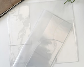 Puede incluir: Porta menús de plástico transparente con la parte superior curvada y un bolsillo. El texto "KOLOUR CAFE" está impreso en la parte delantera. Un diario de cuero marrón con dijes dorados y una ramita de vegetación están en el fondo.