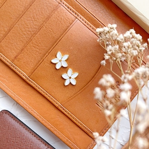 May include: Two white flower-shaped earrings with gold accents on a brown leather background. The earrings are small and delicate.