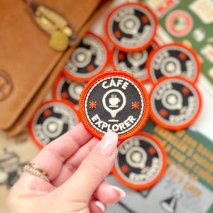 Op de afbeelding: Een verzameling ronde, geborduurde patches met een zwarte achtergrond en een oranje rand. De patches tonen de woorden "CAFE EXPLORER" en een koffiekopje-icoon. Een hand houdt een patch vast, andere liggen verspreid.