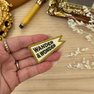 May include: A cream-coloured patch with the words "WANDER & WONDER" in black lettering. The patch has a gold border and a pennant shape. The patch is held in a hand.