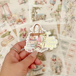 Puede incluir: Una pegatina decorativa con un bolso de compras, libros y cerezas. La pegatina tiene el texto "Now Showing: Your Best Days" en dorado. El fondo presenta otras pegatinas con diseños florales y vintage.