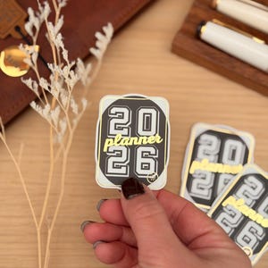 Pode incluir: Uma mão segurando um adesivo com o texto "2026 planner" em um design preto, branco e dourado. O adesivo tem formato retangular com cantos arredondados. Outros adesivos e uma carteira de couro estão ao fundo.
