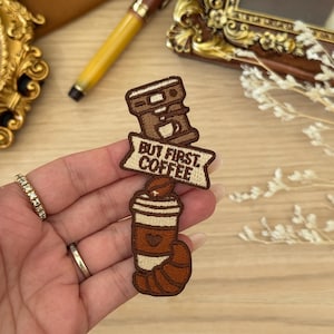 Op de afbeelding: Geborduurde patch met een koffiezetapparaat en een koffiekopje met de tekst "BUT FIRST COFFEE". De patch is bruin en beige met een hartdetail op de beker. Een koffieboon staat bovenop de beker.