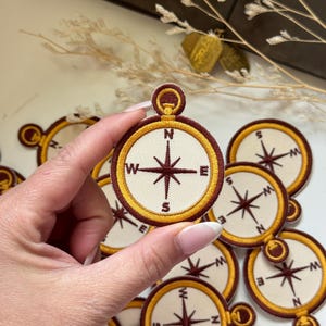 Op de afbeelding: Geborduurde kompaspatches met een crèmekleurige wijzerplaat, een donkerbruine windroos en een goudkleurige rand. De windroos heeft de letters N, S, E en W. De patches zijn geschikt om op kleding of accessoires te naaien.