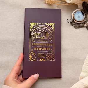 以下が含まれることがあります： 「FOR ALL IN LOVE WITH WORDS」と「ANTHOLOGY OF ADVENTURES MEMORIES」の文字が金色で書かれた、バーガンディ色のジャーナル。ジャーナルには装飾的なボーダーがあり、背景には小さなコンパスがあります。