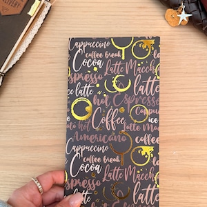 Puede incluir: Un cuaderno gris oscuro con un diseño temático de café. La portada presenta las palabras "Cappuccino", "Latte" y "Espresso" en una escritura rosa, junto con manchas doradas de tazas de café. El cuaderno está en manos de una persona.