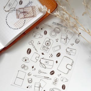 Pode incluir: Uma folha de autocolantes com ilustrações de café, incluindo grãos de café, chávenas e carteiras. Os autocolantes são a preto e branco, com o texto "KAFE KORNER VOL." e "KOLOUR CAFE & STABLEPLANS". Um caderno de couro castanho está no fundo.