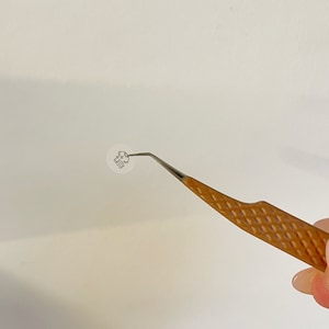 May include: A pair of tweezers with a brown, textured handle and a silver metal tip. The tweezers are holding a small, white, round sticker with a black design.