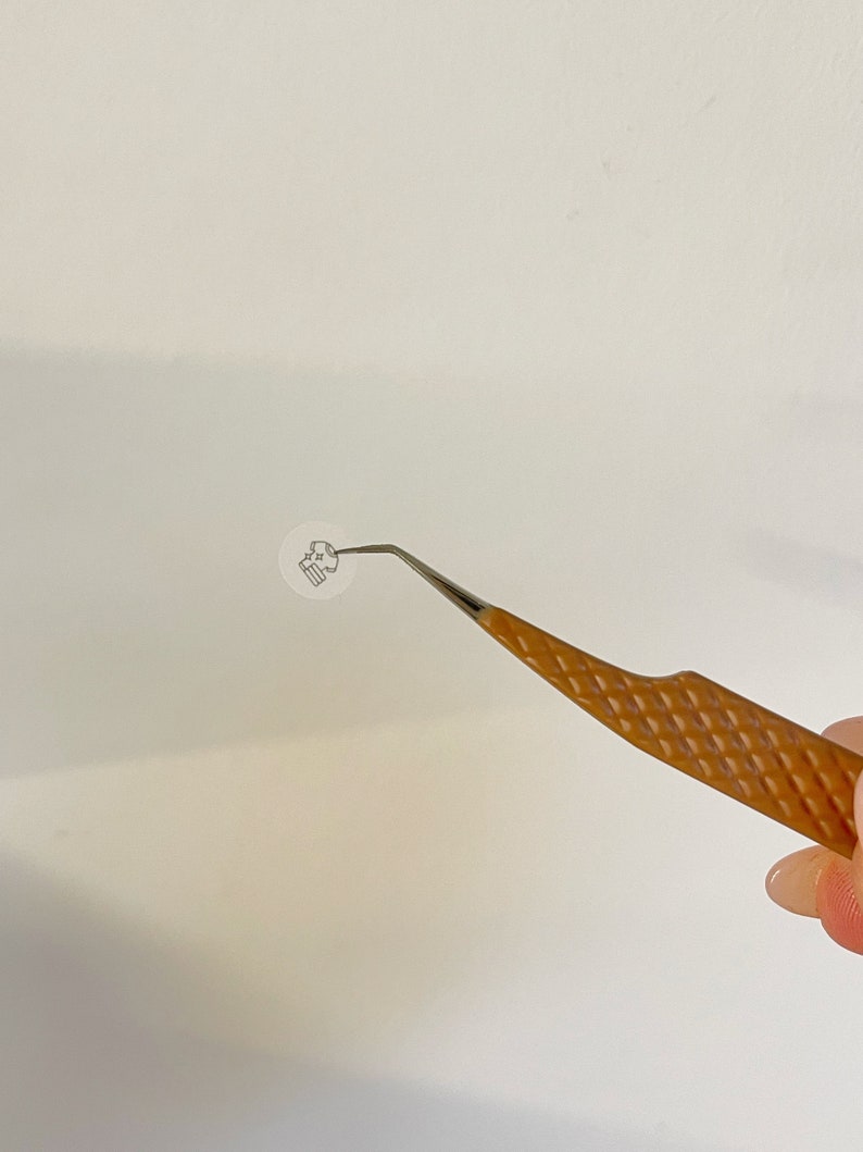 May include: A pair of tweezers with a brown, textured handle and a silver metal tip. The tweezers are holding a small, white, round sticker with a black design.