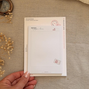 Könnte beinhalten: Ein weißer, rechteckiger Notizblock mit schwarzem Aufdruck, darunter 'FROM THE DESK OF a paper lover', 'DATE', 'LOCATION' und ein rotes Stempelmotiv. Der Block hat im unteren Bereich horizontale Linien und eine kleine rote Ticketgrafik.