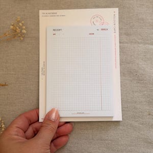 May include: A cream-colored notepad with a receipt-style grid design. The top sheet features the word RECEIPT, a date and location line, and a grid pattern with a vertical red line. The header includes the text KOLOUR CAFE and a small stamp graphic.