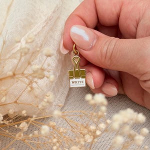 May include: A gold-toned metal charm shaped like a binder clip with a white enamel base featuring the word WRITE printed in black capital letters. The charm includes a small circular jump ring at the top for attachment to a chain or bracelet.
