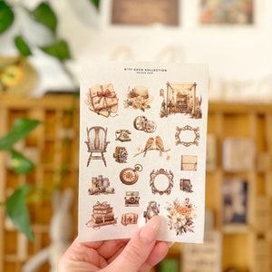 May include: A sheet of vintage-style stickers featuring images of books, cameras, birds, a rocking chair, a clock, and a floral arrangement. The stickers are labeled "N°77 Deco Collection - Colour Cafe".