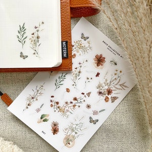 May include: A sheet of stickers featuring a floral design with butterflies. The stickers are on a white background and have a rustic, vintage aesthetic. The text "DECO KOLLECTION N&deg;1" and "KOLOUR CAFE" is printed on the sheet.