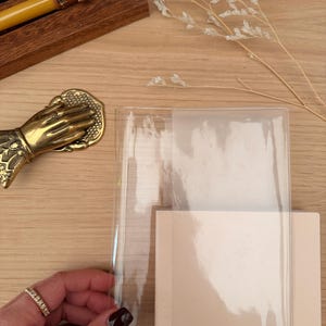 以下が含まれることがあります： 透明なプラスチックカバーが付いた、明るいベージュ色のノート。指輪をした手がカバーを持っています。背景には、いくつかのペンが入った木製のペンホルダーがあります。装飾的な真鍮の手も見えます。