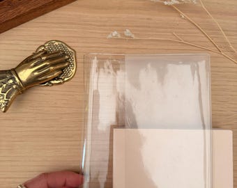 Puede incluir: Un cuaderno beige claro con una cubierta de plástico transparente. Una mano con un anillo sostiene la cubierta. Un portalápices de madera con varios bolígrafos está en el fondo. También se ve una mano decorativa de latón.