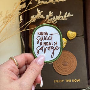 Peut inclure: Un patch brodé blanc avec des coutures vertes. Le patch dit "Kinda sweet kinda savage" en broderie brune.