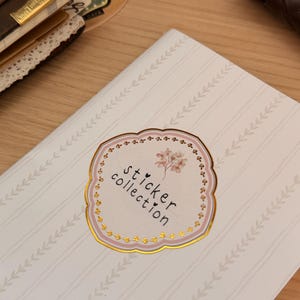 May include: A notebook with a light pink cover featuring a pattern of vertical stripes and small leaf designs. The cover has a sticker that says "sticker collection" in a decorative font, with a floral illustration.