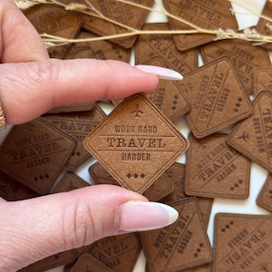 Puede incluir: Primer plano de una mano sosteniendo un parche de cuero marrón con el texto "Work Hard Travel Harder" y un pequeño icono de avión. El parche está rodeado de otros parches similares con el mismo texto.