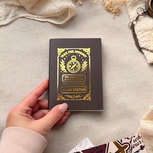 Może przedstawiać: Mały, ciemnobrązowy notatnik ze złotymi literami i wzorem kompasu. Tekst brzmi "TAKE THE JOURNEY" i "ATLAS STATION". Notatnik trzymany jest w dłoni, a pod spodem widoczne są naklejki o tematyce podróżniczej.