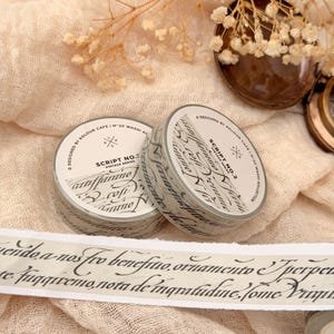 May include: Two rolls of washi tape with script lettering, labelled "Script No. 2 Vintage Series." A strip of tape with script text is unrolled. The tape is light grey with black lettering, set against a beige fabric background.