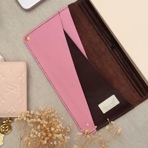 Può includere: Una copertina per diario in pelle rosa e marrone con un taccuino color crema all'interno. La copertina ha una tasca ed è fissata con elastici. Sono visibili rivetti color oro. Un portafoglio rosa chiaro e un portachiavi dorato sono sullo sfondo.