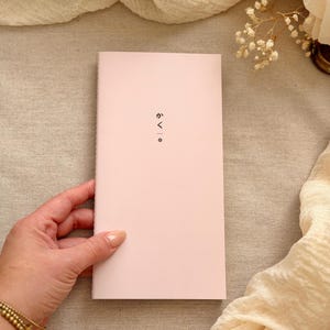 May include: A rectangular, light pink notebook with a vertical orientation. The front cover features the Japanese text "かく" followed by a vertical line and a small flower icon printed in black ink. The notebook has a clean, simple design.