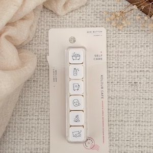 May include: A set of six white square mini button stamps on a rectangular backing card labelled MINI BUTTON and SELF CARE. Each stamp features a black line drawing of a self-care activity, including a bathtub, lotion, journal, face mask, yoga, and sleep.
