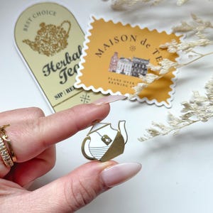 May include: Gold teapot enamel pin with a white tea bag hanging out of the spout. The pin is being held by a hand with a gold ring on the finger. The background is a white surface with two stickers, one that says "Best Choice Herbal Tea" and the other that says "Maison de Te Planted Over Brewing"