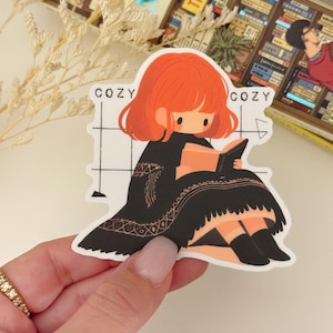 May include: A sticker featuring a cartoon illustration of a girl with red hair sitting in a chair and reading a book. The sticker has a white background with black and white grid lines. The text "Cozy" appears twice on the sticker.