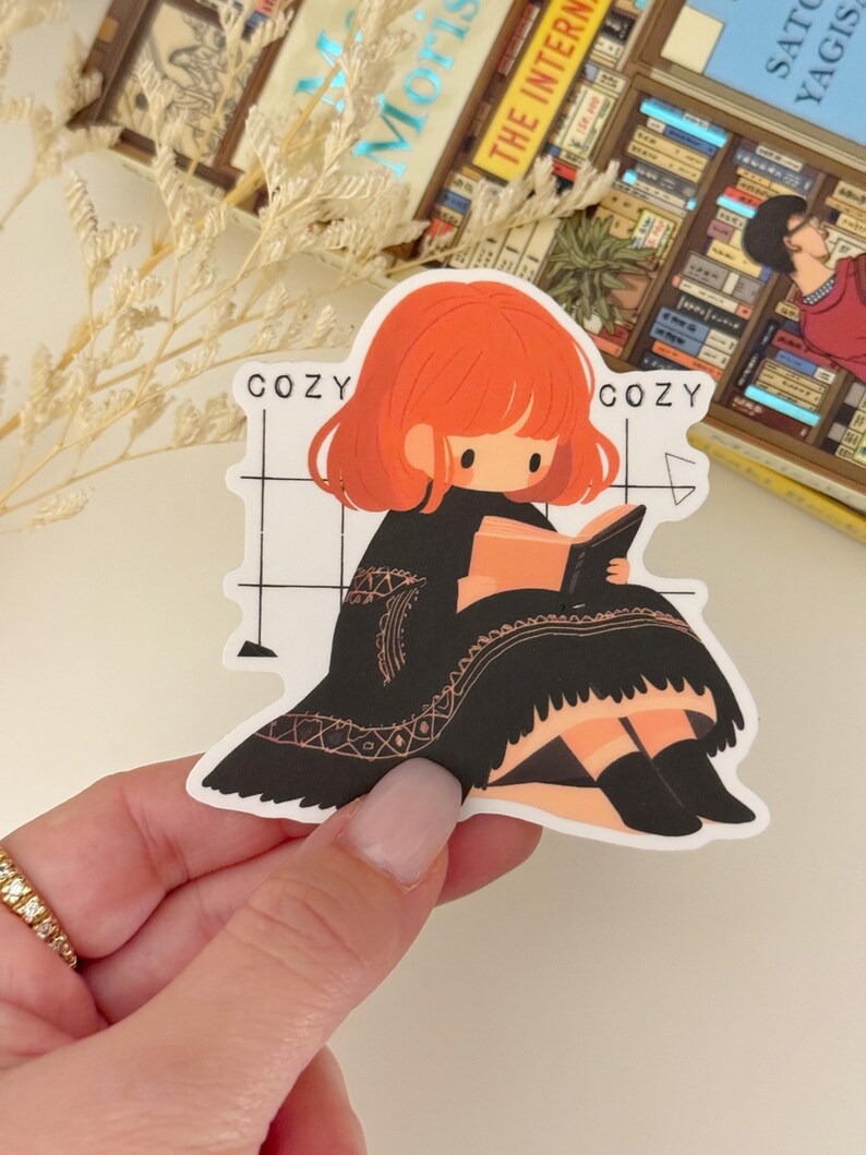 May include: A sticker featuring a cartoon illustration of a girl with red hair sitting in a chair and reading a book. The sticker has a white background with black and white grid lines. The text "Cozy" appears twice on the sticker.