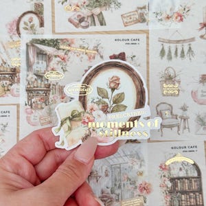 May include: A sticker featuring a rose illustration within a circular frame, with the words "PRESCRIBED: moments of stillness" in gold. The background shows other stickers with floral and apothecary designs, and the sticker is held in a hand.