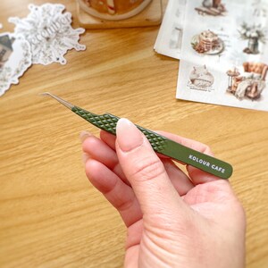 May include: Green tweezers with a slanted tip and a textured grip. The tweezers have the words "KOLOUR CAFE" printed on the side.