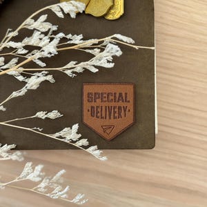 Op de afbeelding: Een donkerbruin leren notitieboek met een patch met de tekst "SPECIAL DELIVERY". Het notitieboek heeft een zwarte elastische sluiting en gouden bedels. Witte droogbloemen staan op de voorgrond, op een houten oppervlak.
