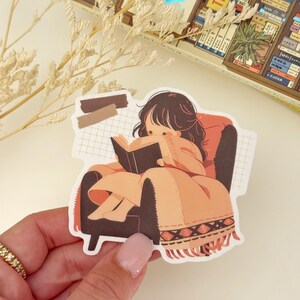 May include: A sticker featuring a cartoon illustration of a person sitting in a chair and reading a book. The person has brown hair and is wearing a brown dress. The chair is brown and has a patterned blanket on it. The background is a grid pattern.