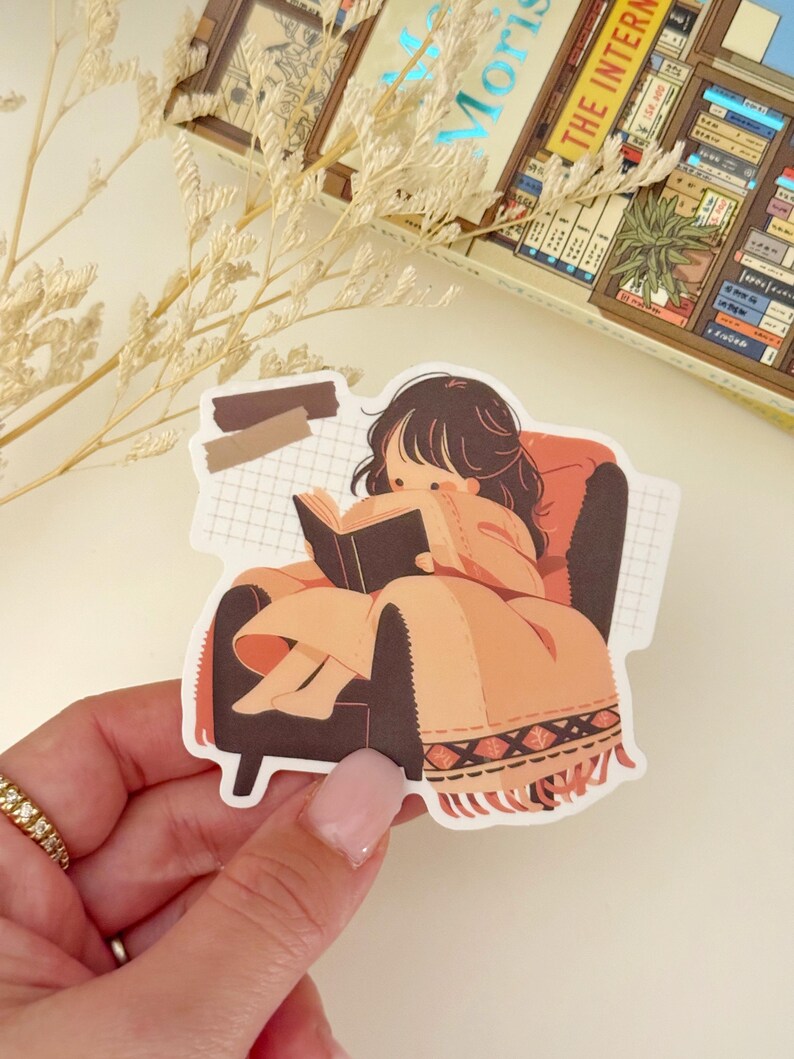 May include: A sticker featuring a cartoon illustration of a person sitting in a chair and reading a book. The person has brown hair and is wearing a brown dress. The chair is brown and has a patterned blanket on it. The background is a grid pattern.