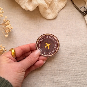 May include: A circular sticker with a brown border and the words "LOVE, LUGGAGES, AND LATE FLIGHTS". A yellow airplane graphic is over a world map. The sticker is held by a hand with a gold ring.