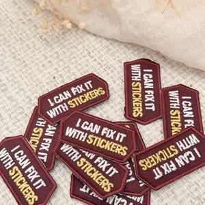 May include: A collection of small, rectangular embroidered patches in a deep maroon color. Each patch features a dark maroon border and the text "I CAN FIX IT WITH STICKERS" embroidered in white and yellow thread, with the word STICKERS in yellow.