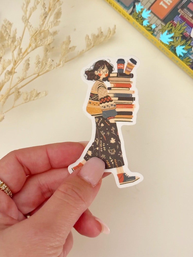 May include: A sticker of a cartoon woman wearing a brown and white sweater and a black skirt with a floral pattern. She is carrying a stack of books and two coffee cups.