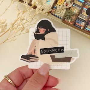 May include: A sticker featuring a cartoon illustration of a person reading a book while wrapped in a blanket. The sticker has a white background with a grid pattern and the text "BOOKWORM" in black.