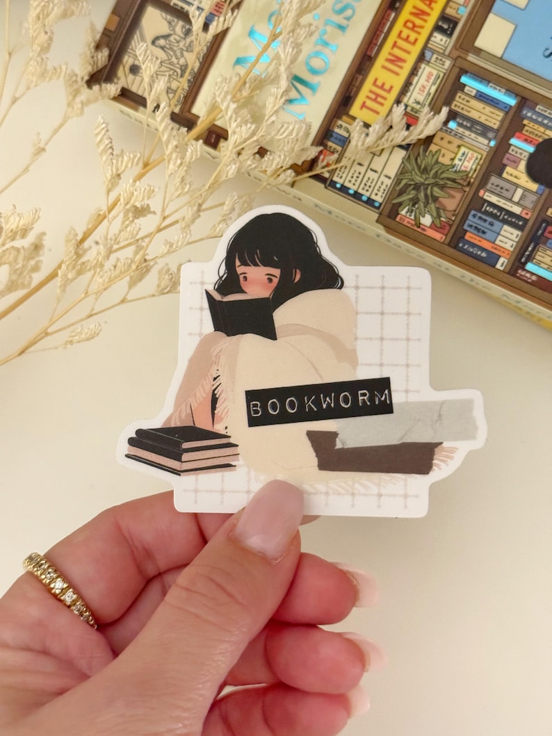 May include: A sticker featuring a cartoon illustration of a person reading a book while wrapped in a blanket. The sticker has a white background with a grid pattern and the text "BOOKWORM" in black.